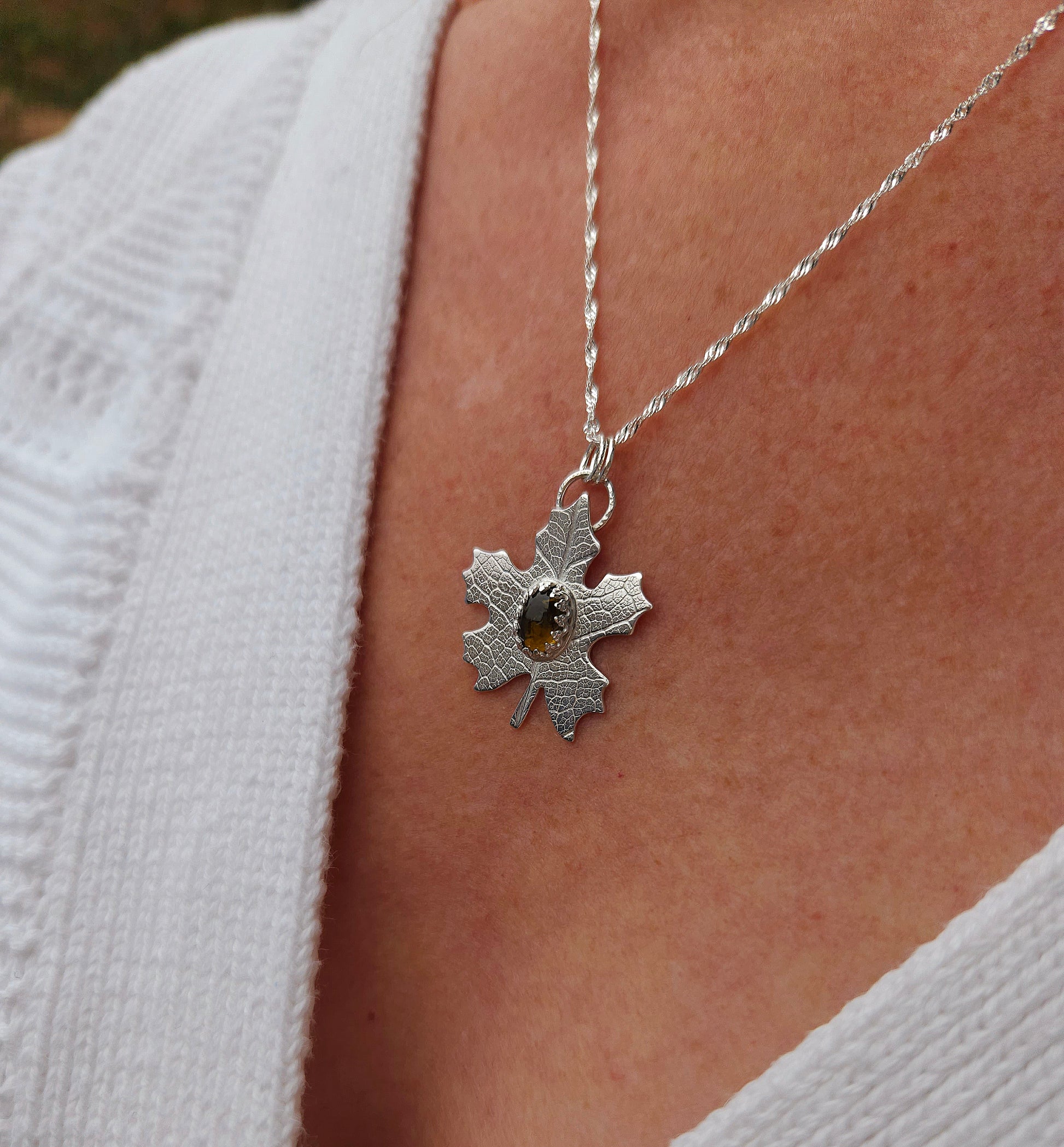 A sterling silver leaf pendant with brown tourmaline gemstone being worn with a white cardigan.