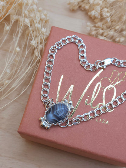 A silver chain bracelet with a Botswana Agate gemstone charm, displayed on a gift box with dried flowers in the background.