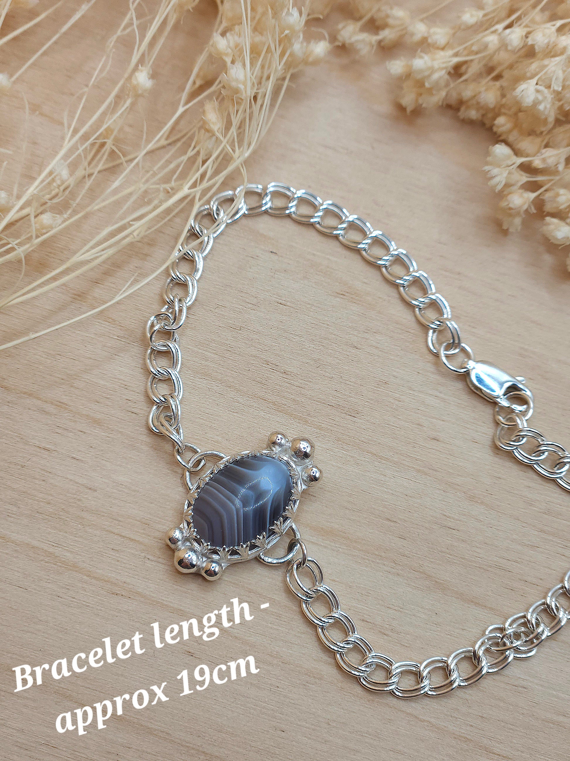 A silver chain bracelet with Botswan agate gemstone displayed on a wooden background with dried flowers. A note in the bottom left corner reads 'Bracelet length - approx 19cm'.