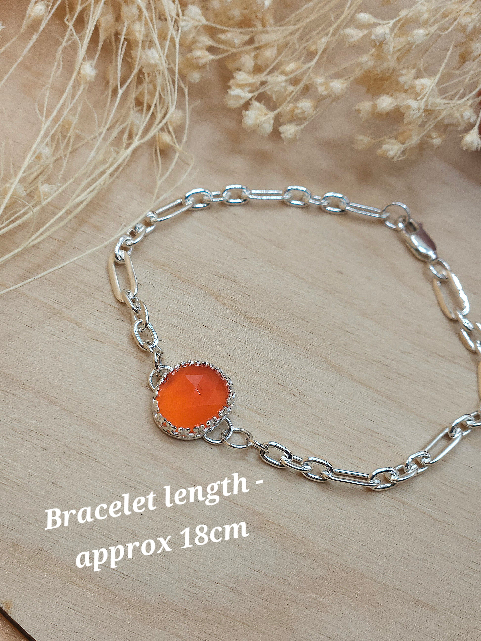 A silver link chain bracelet with orange carnelian gemstone displayed on a wooden background with dried flowers. Wording at the bottom left corner reads 'Bracelet length - approx 18cm'.