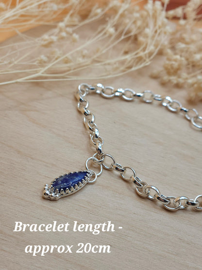 A sterling silver chain bracelet with a navy blue coloured Lapis Lazuli gemstone charm. Displayed on a wooden background with dried flowers. A note in the left bottom corner of the photo reads 'Bracelet length - approx 20cm'.