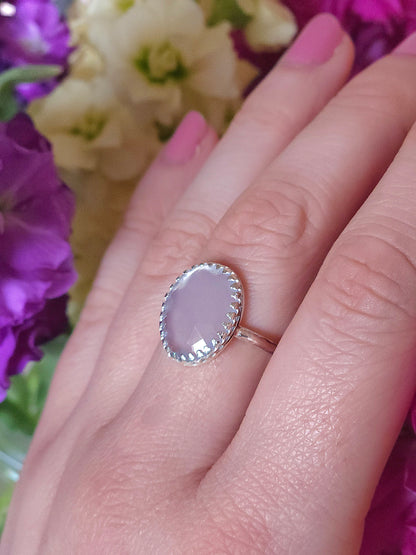A hand modelling a silver ring with a lilac coloured chalcedony gemstone. Pink, yellow and purple flowers in the background.