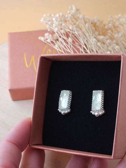 A hand holding an open gift box displaying a pair of silver stud earrings with green prehnite gemstones.