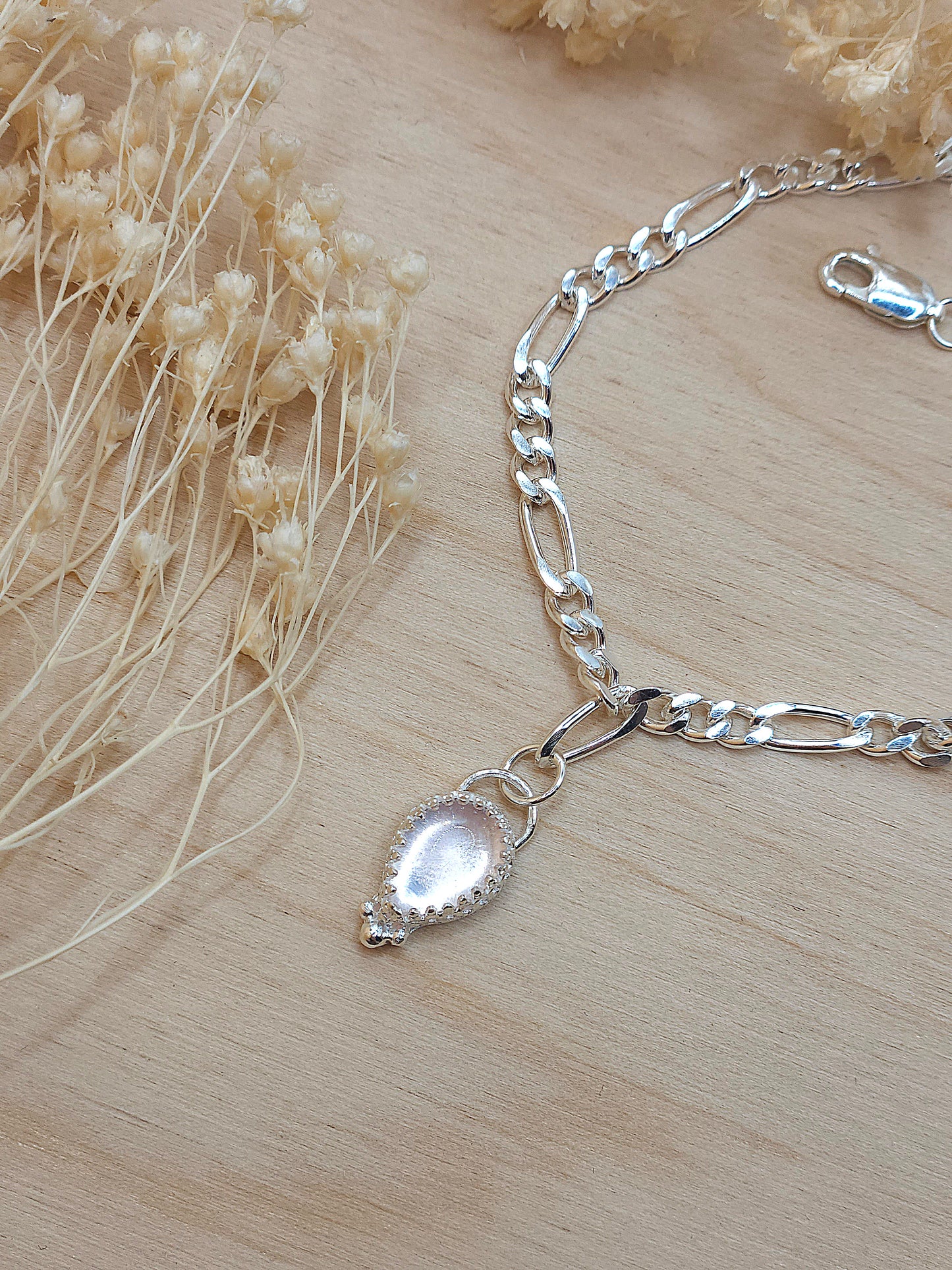 A sterling silver chain bracelet with rose quartz gemstone displayed on a wooden background with dried flowers surrounding it.