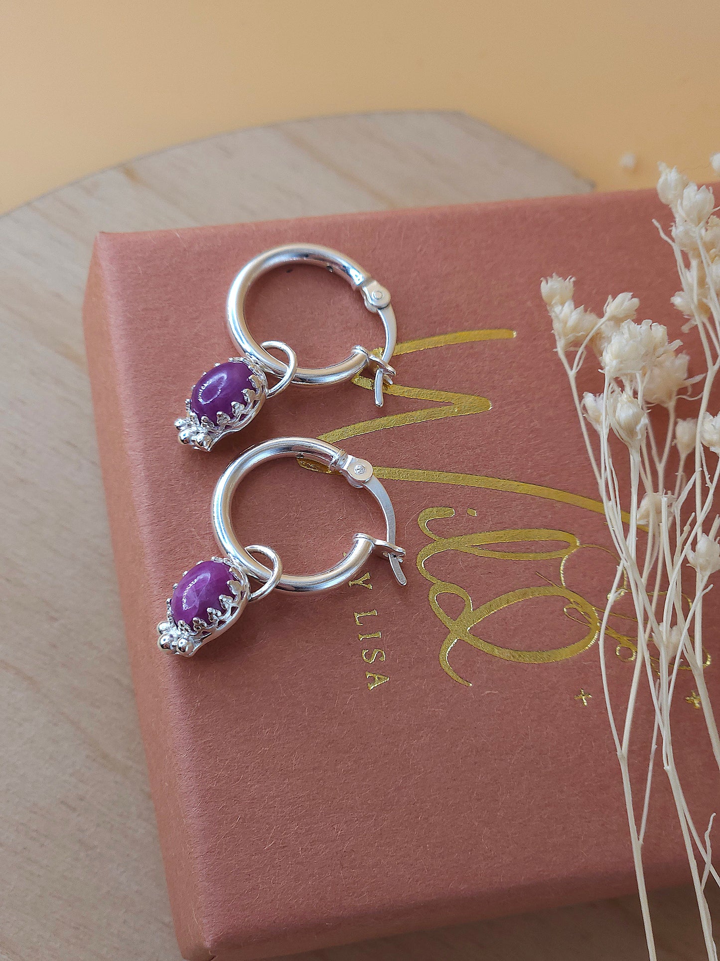 A pair of silver hoop earrings with ruby gemstones lay on a terracotta and gold coloured gift box