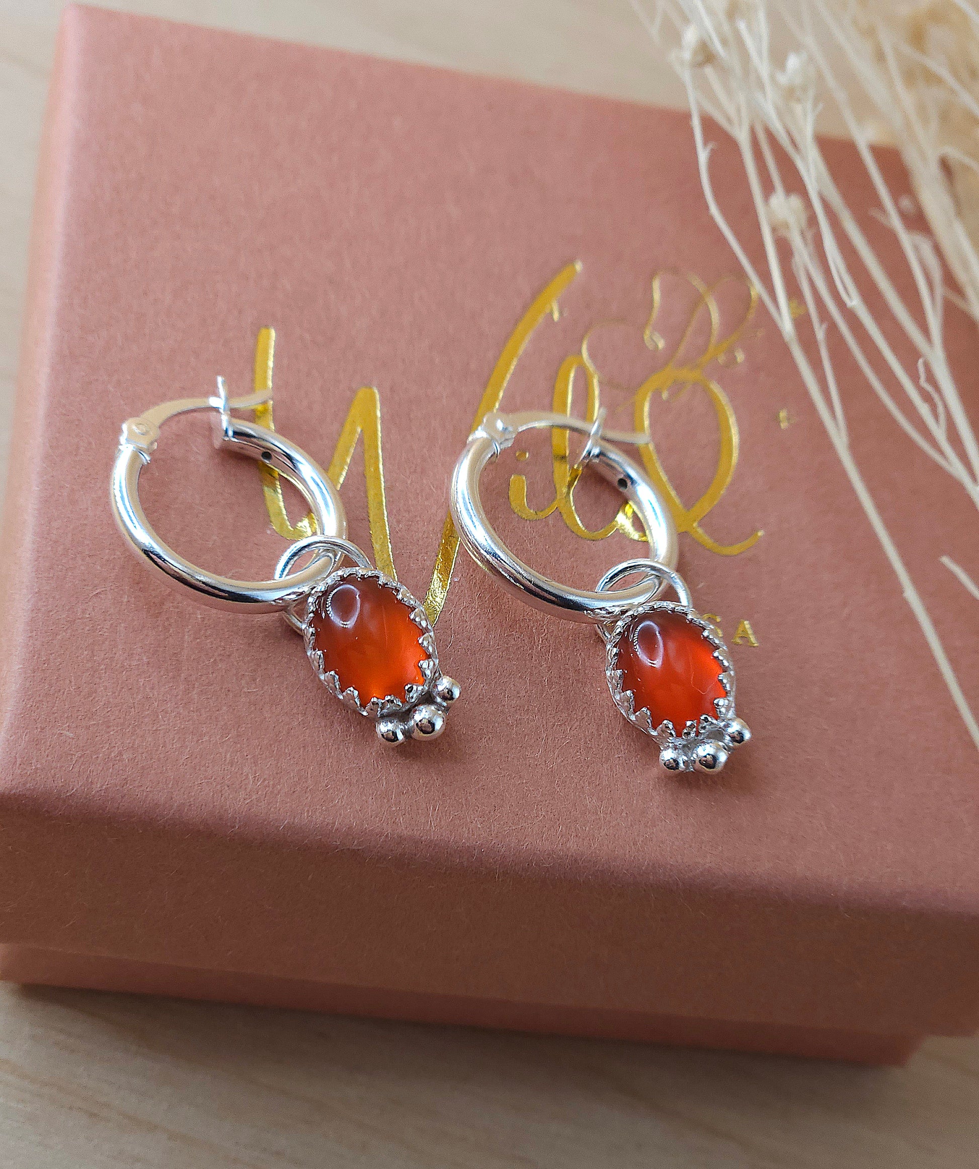 A pair of silver mini hoop earrings with red carnelian charms, displayed on a terracotta and gold-coloured box with a dried flowers.