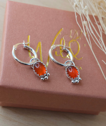 A pair of silver mini hoop earrings with red carnelian charms, displayed on a terracotta and gold-coloured box with a dried flowers.