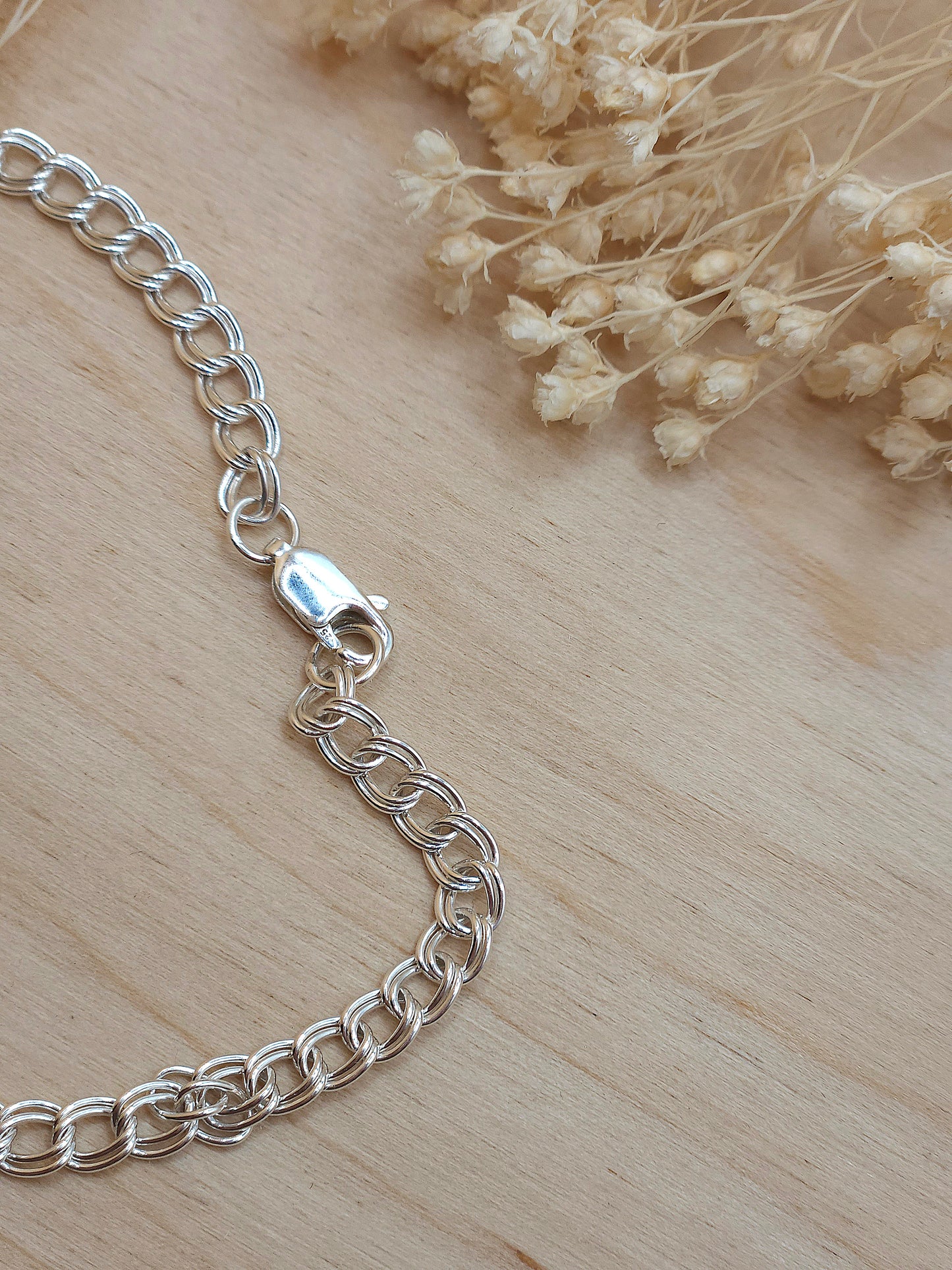 A sterling silver chain bracelet displayed on a wooden background with dried flowers, showing the lobster clasp.