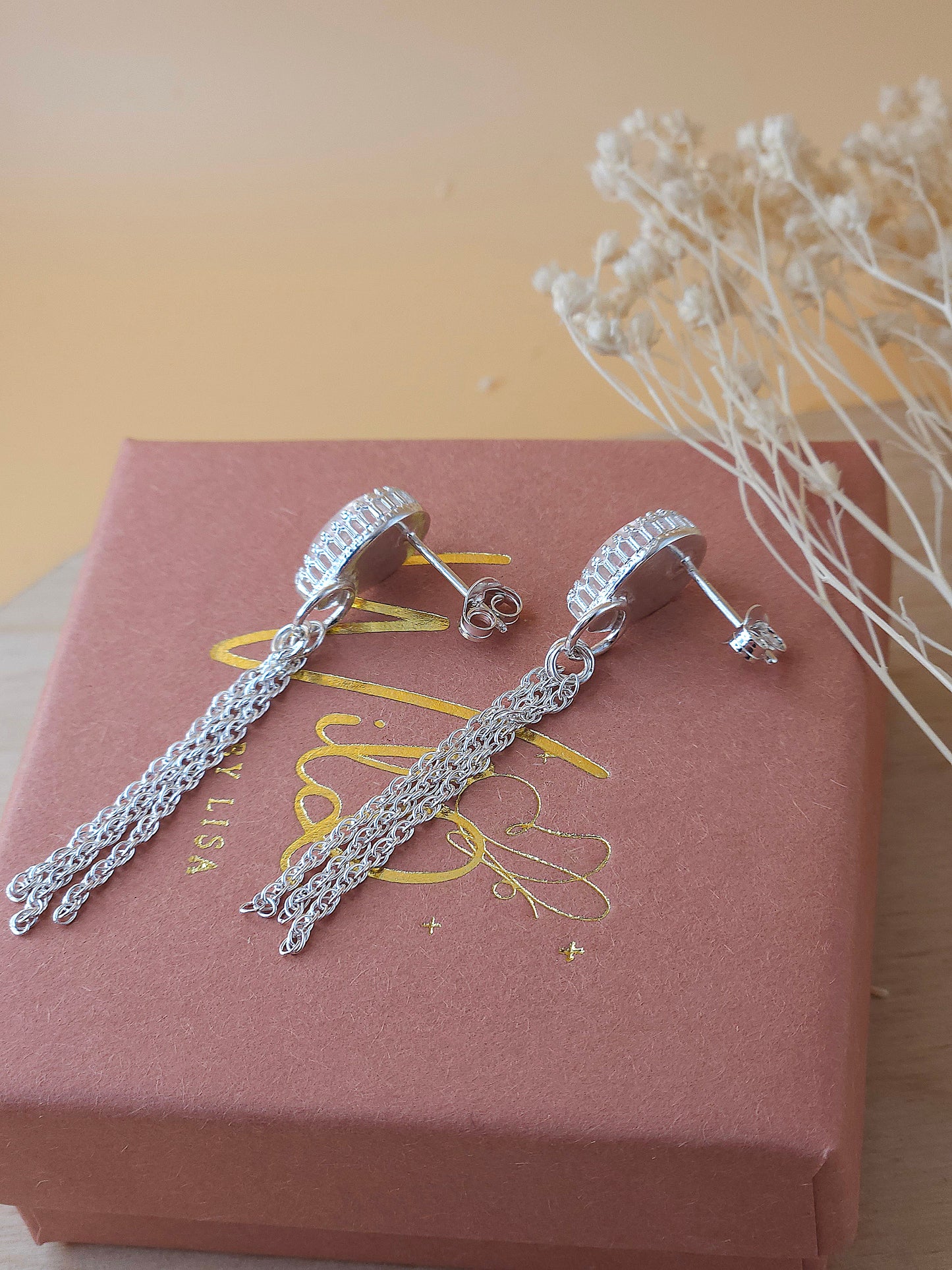 A pair of silver chain dangle earrings lay on a gift box showing the side of the earrings with dried flowers in the background.