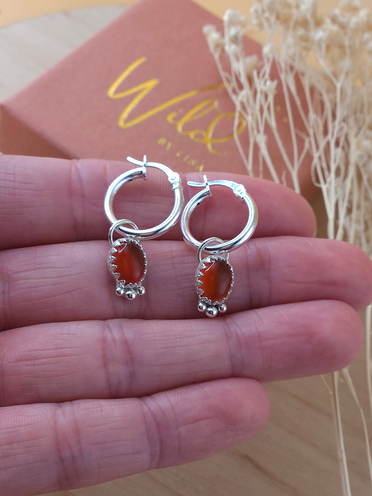 A hand holding a pair of silver hoop earrings with red carnelian charms.