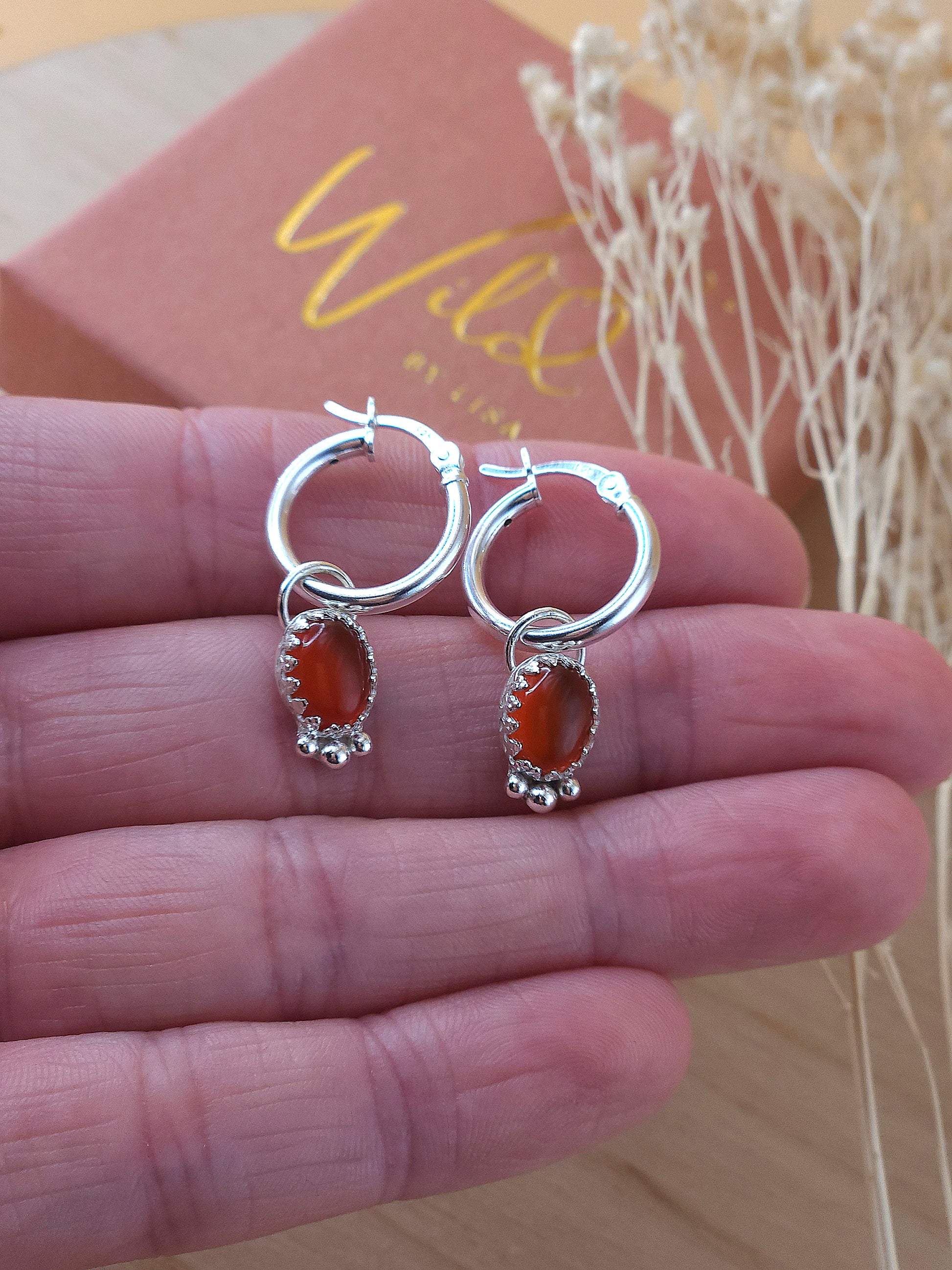 A hand holding a pair of silver hoop earrings with red carnelian charms.