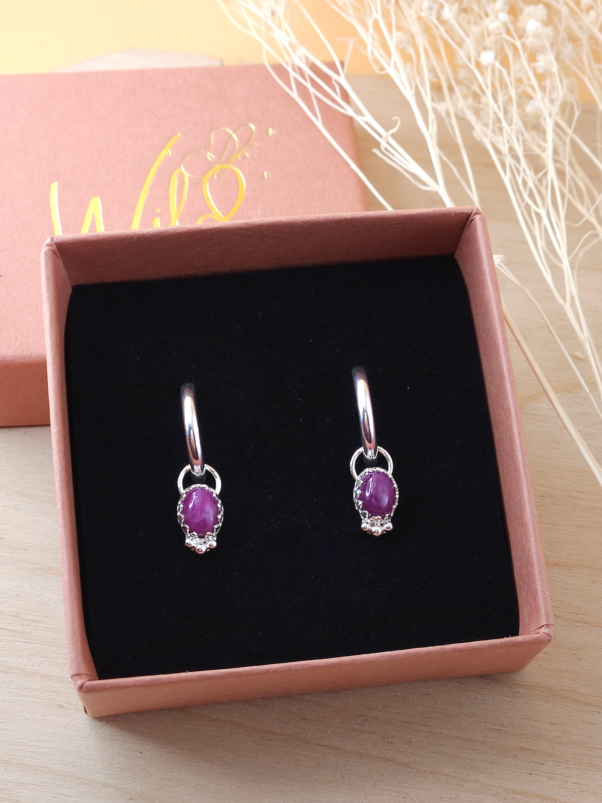 A pair of mini silver hoops with ruby gemstone charms displayed in an open gift box on the black foam insert.