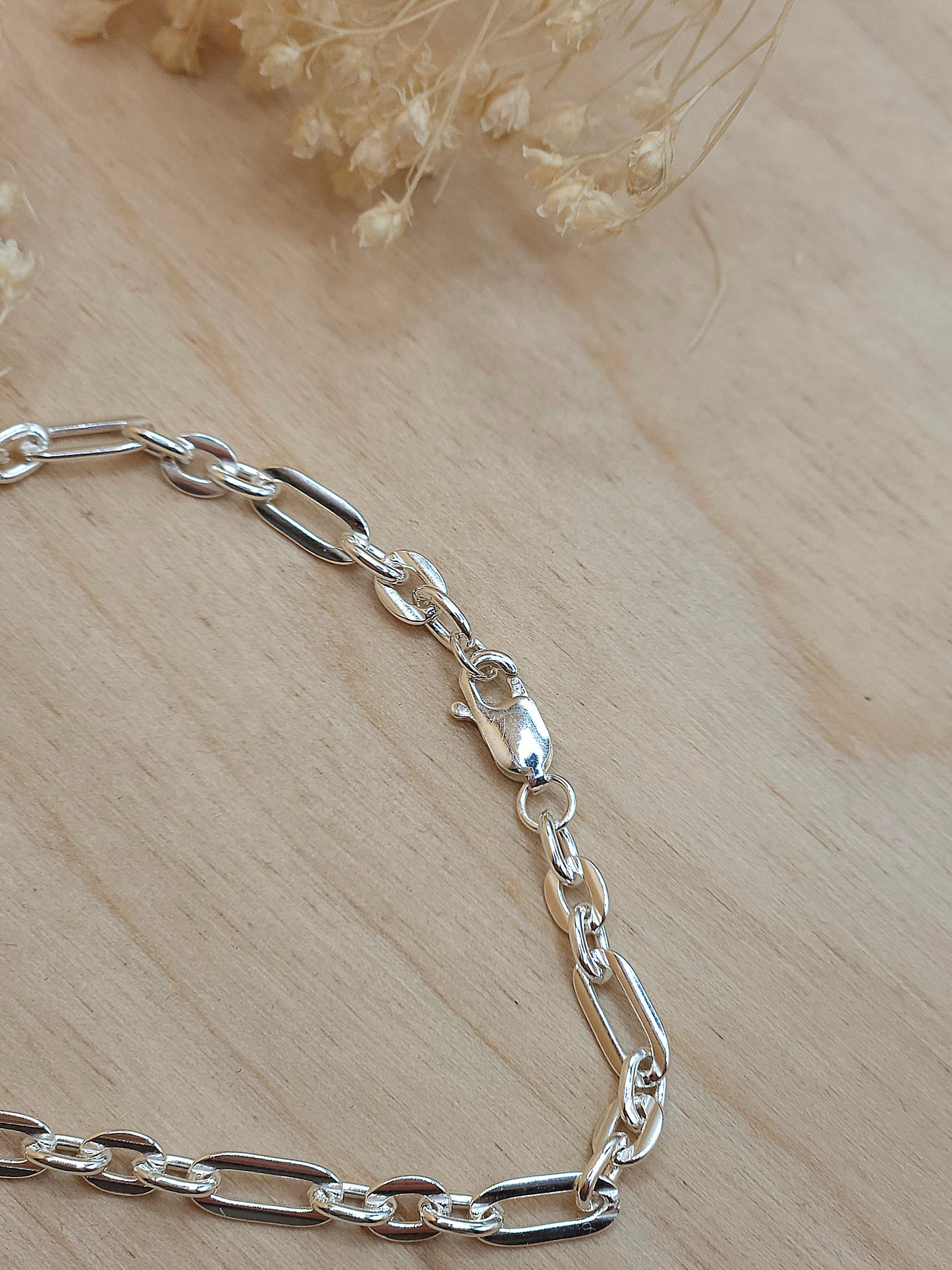 A sterling silver link chain bracelet with lobster clasp displayed on a wooden background with dried flowers.