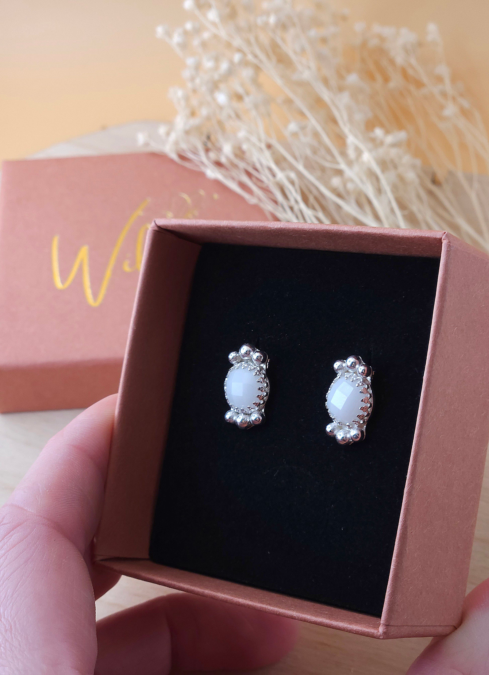 A hand holding an open gift box displaying a pair of white agate gemstone stud earrings with silver embellishments.