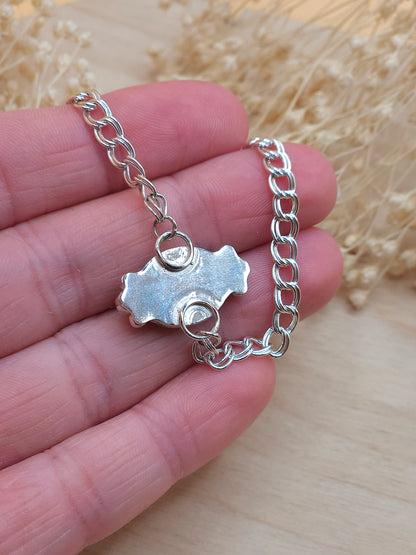 A hand holding a sterling silver chain bracelet, show the back of a gemstone setting. Dried flowers in the background.