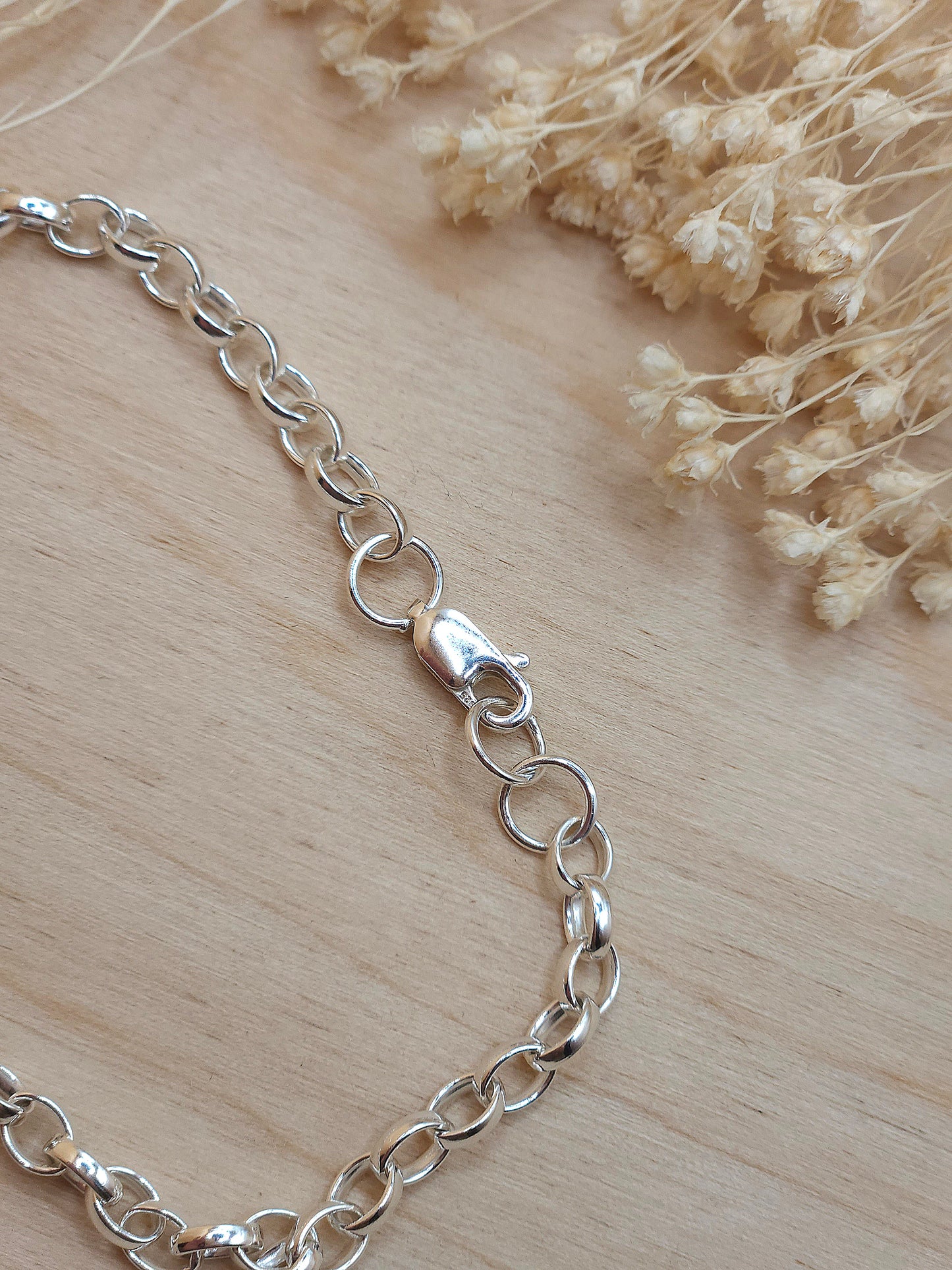 A silver chain bracelet displayed on a wooden background with dried flowers, showing the lobster clasp.