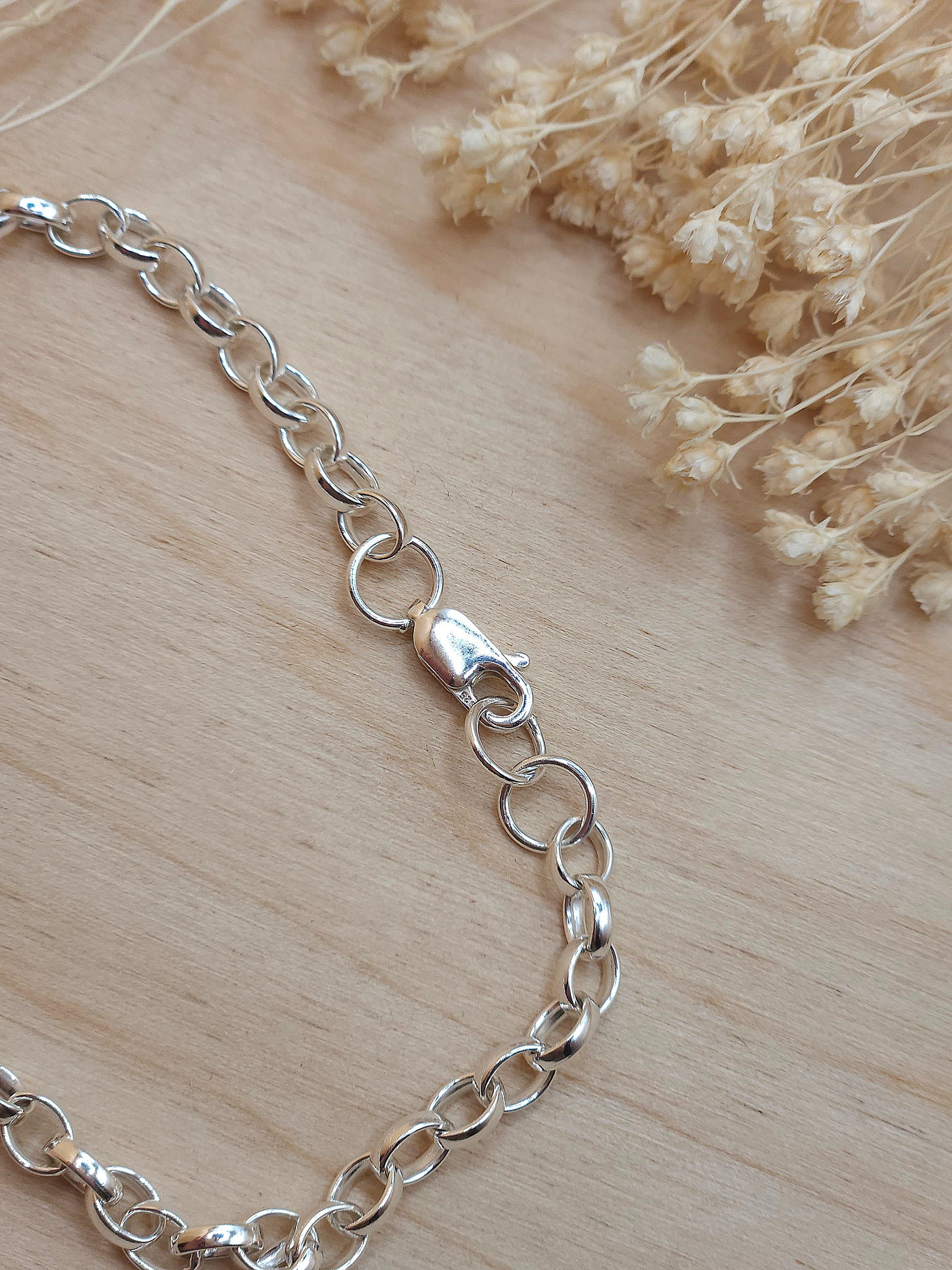 A silver chain bracelet displayed on a wooden background with dried flowers, showing the lobster clasp.