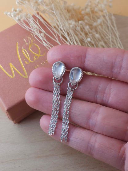 A pair of silver chain dangle earrings with rose quartz gemstones held in a hand, with a small gift box and dried flowers in the background.