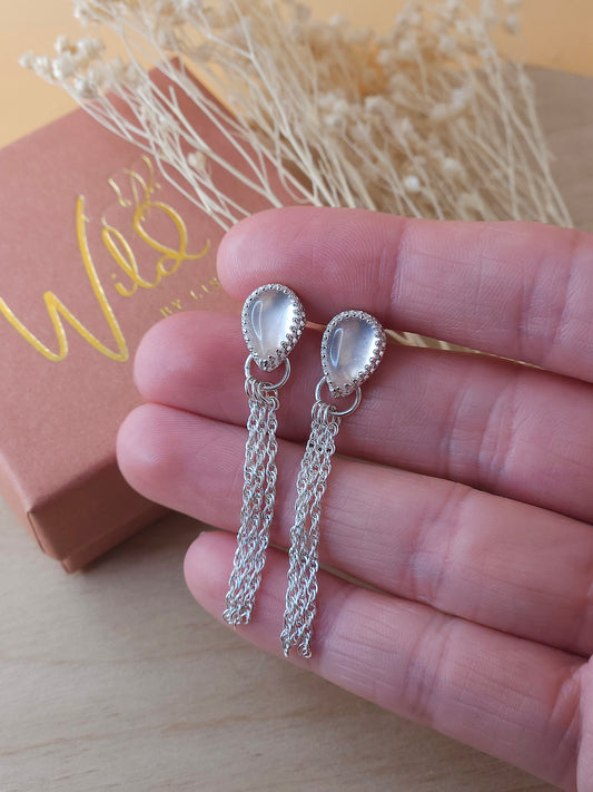 A pair of silver chain dangle earrings with rose quartz gemstones held in a hand, with a small gift box and dried flowers in the background.