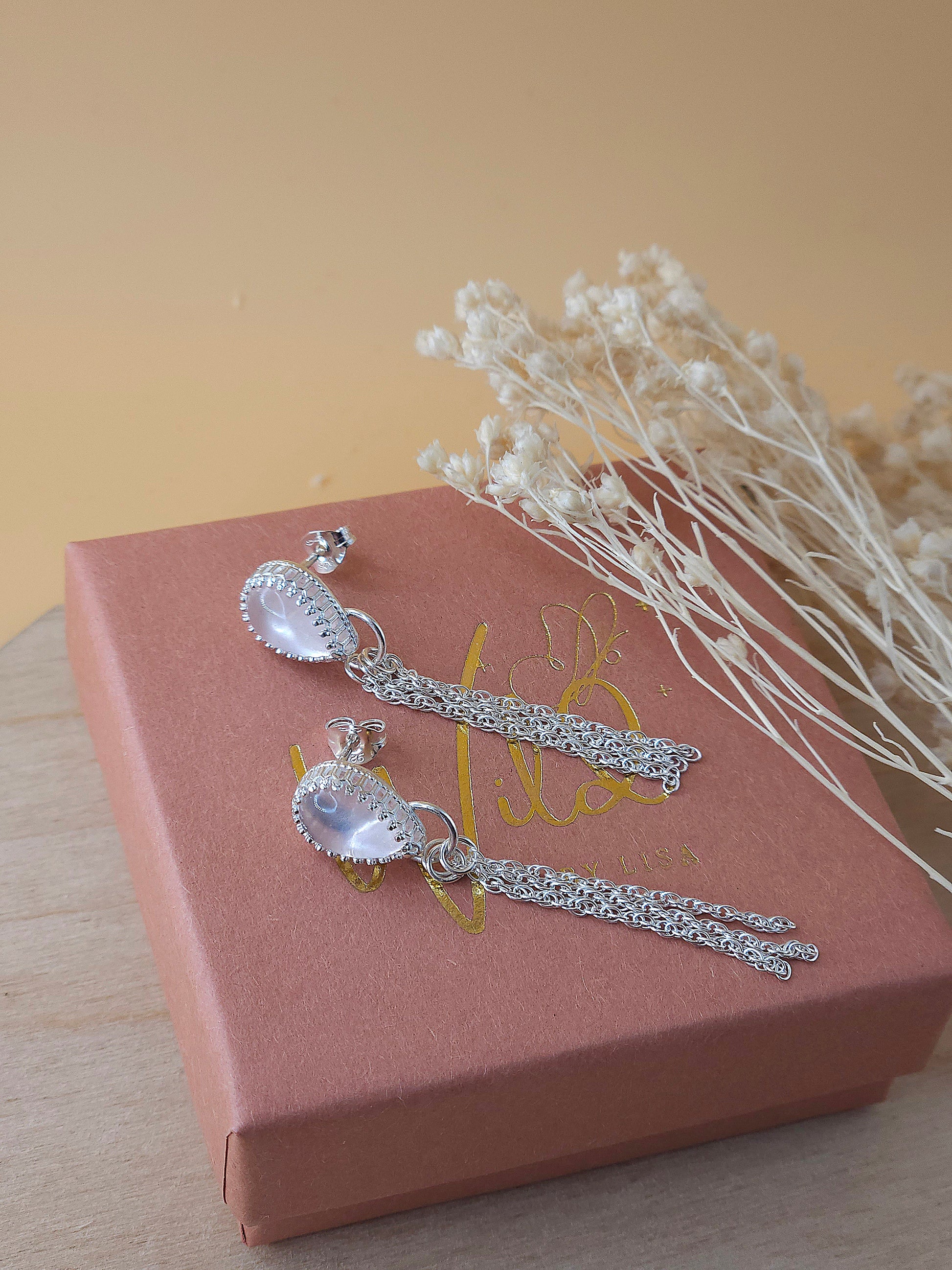 A pair of silver chain dangle earrings with rose quartz gemstones laying on a gift box with dried flowers in the background.