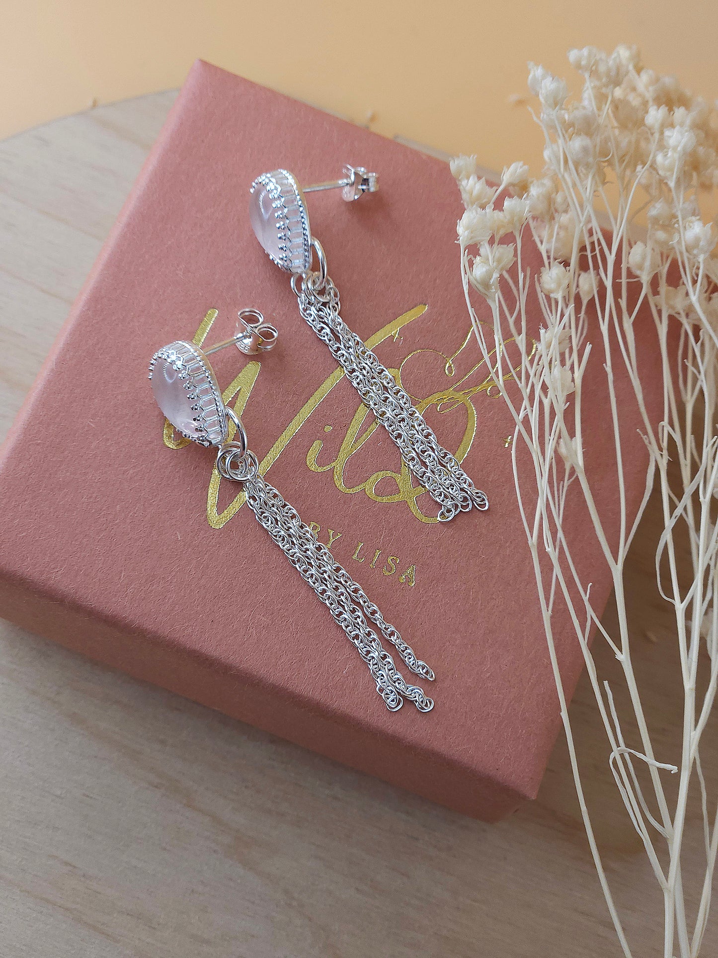 A pair of sterling silver chain earrings with rose quartz gemstones lay on a gift box with dried flowers.