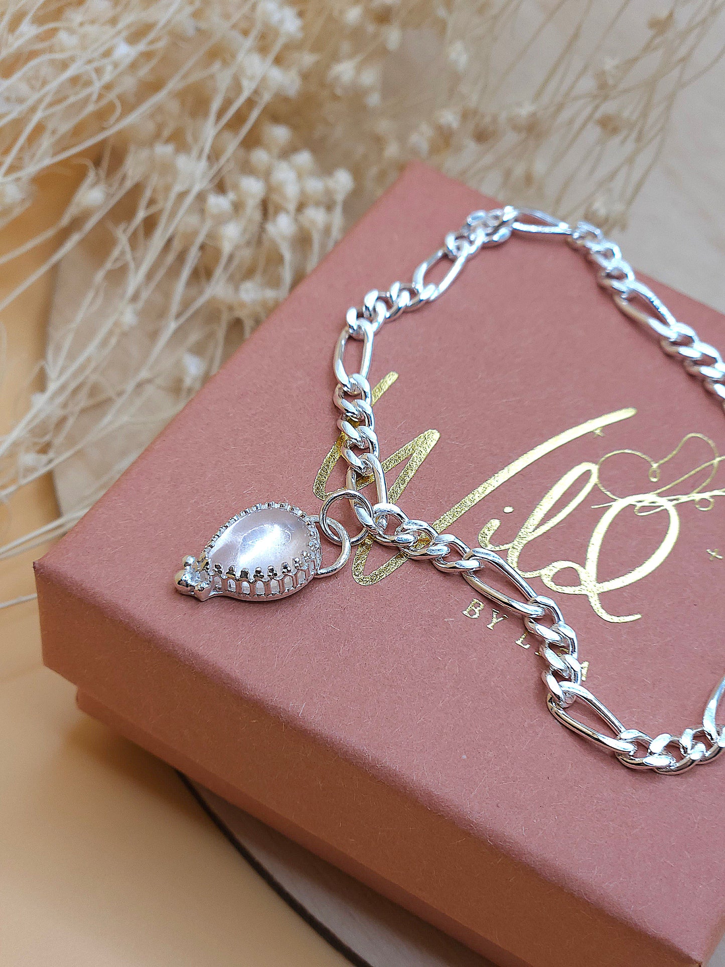 A silver chain bracelet with a rose quartz gemstone charm displayed on  a gift box with dried flowers in the background.