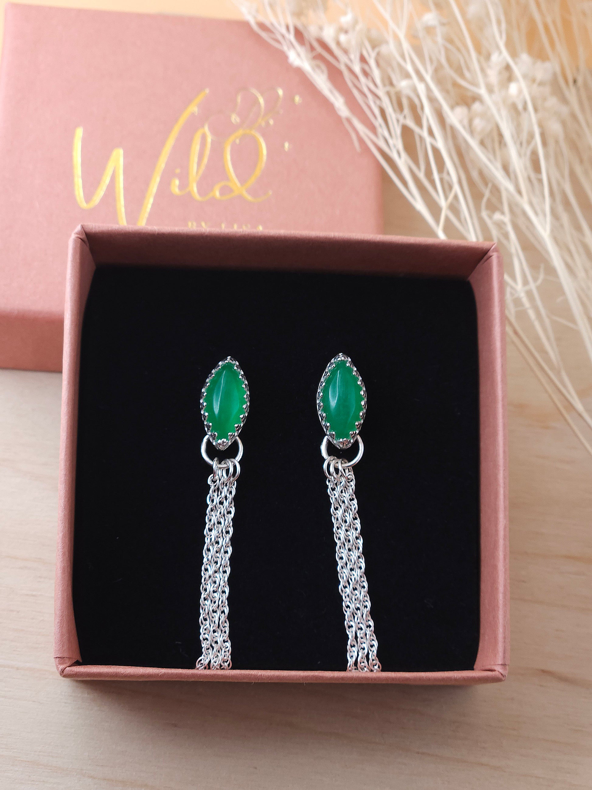 A pair of green aventurine gemstone dangle earrings displayed in an open gift box on a black foam insert with dried flowers in the background.