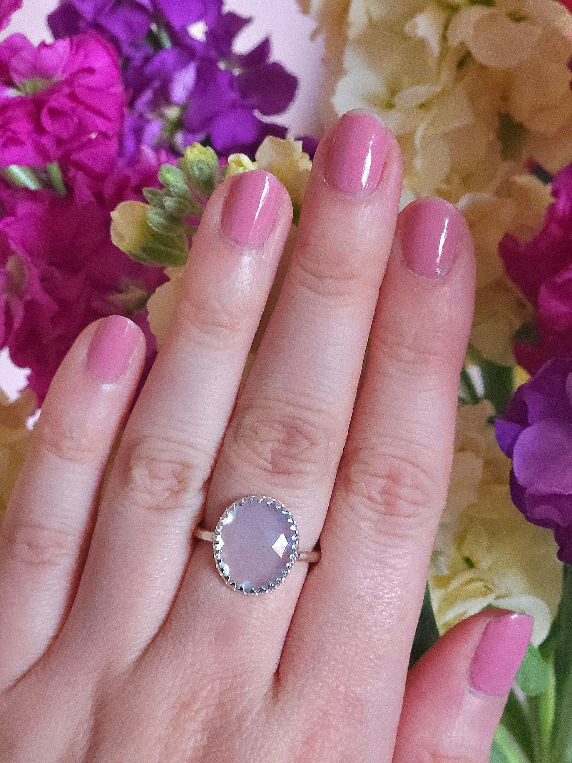 A hand modelling a sterling silver lilac coloured chalcedony gemstone ring with a hidden flower detail. Pink, purple and yellow flowers in the background.