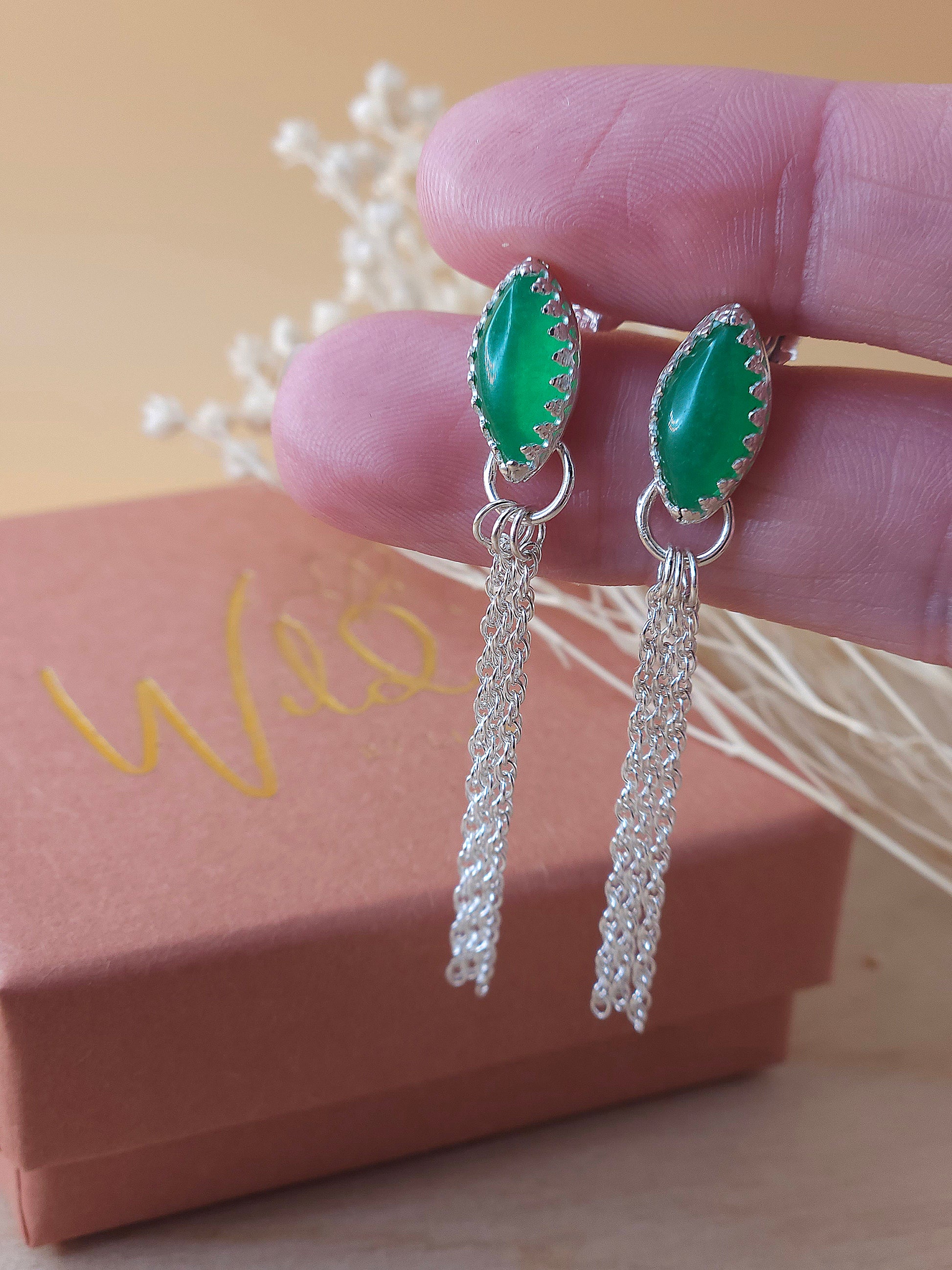 A pair of silver chain dangle earrings with green aventurine gemstones held in a hand, with a small gift box and dry flowers in the background.