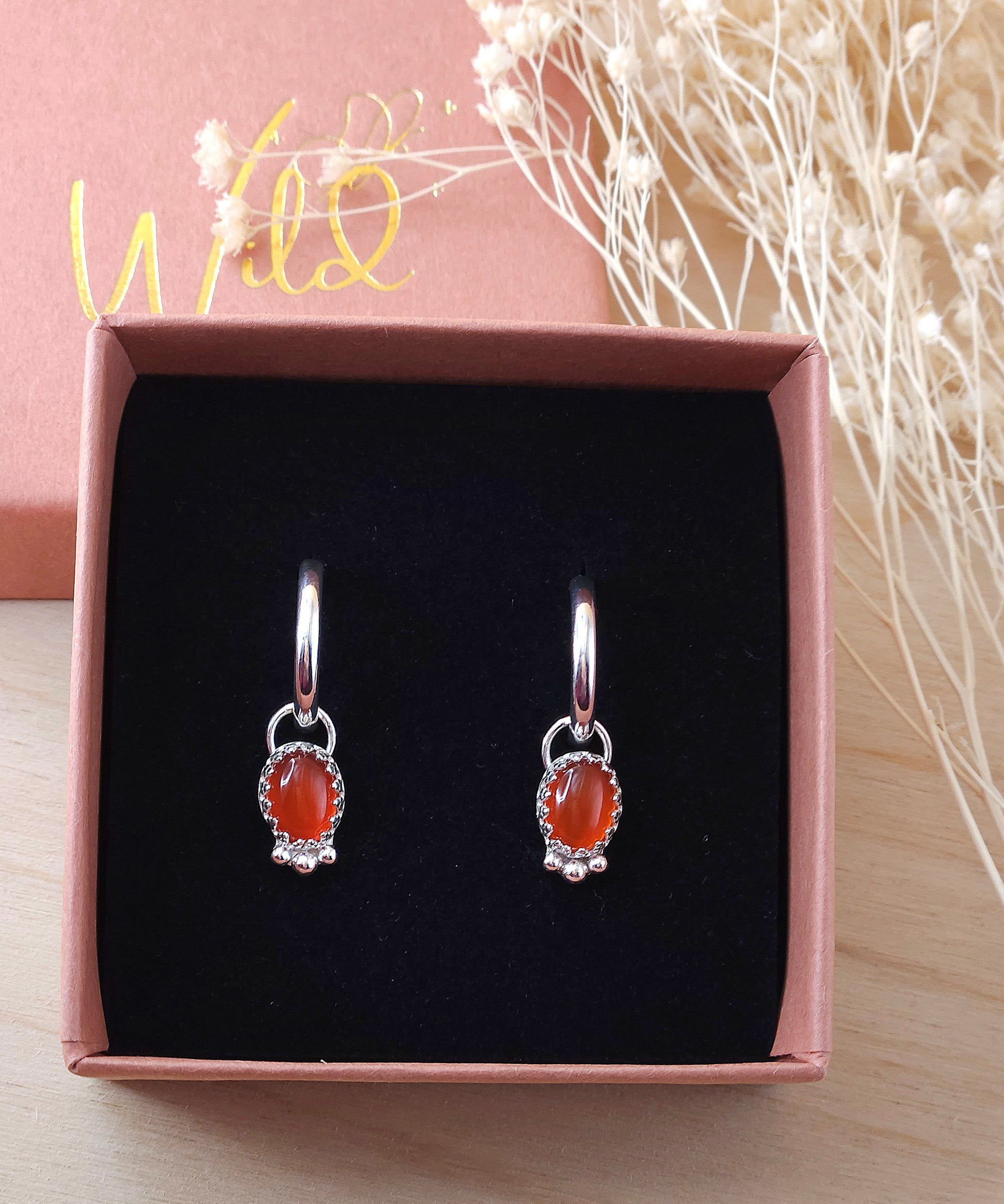 A pair of silver hoop earrings with red carnelian gemstones displayed in an open gift box with black foam insert.