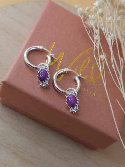 A pair of silver mini hoop earrings with a ruby charm, displayed on a terracotta and gold coloured gift box with decorative dried flowers.