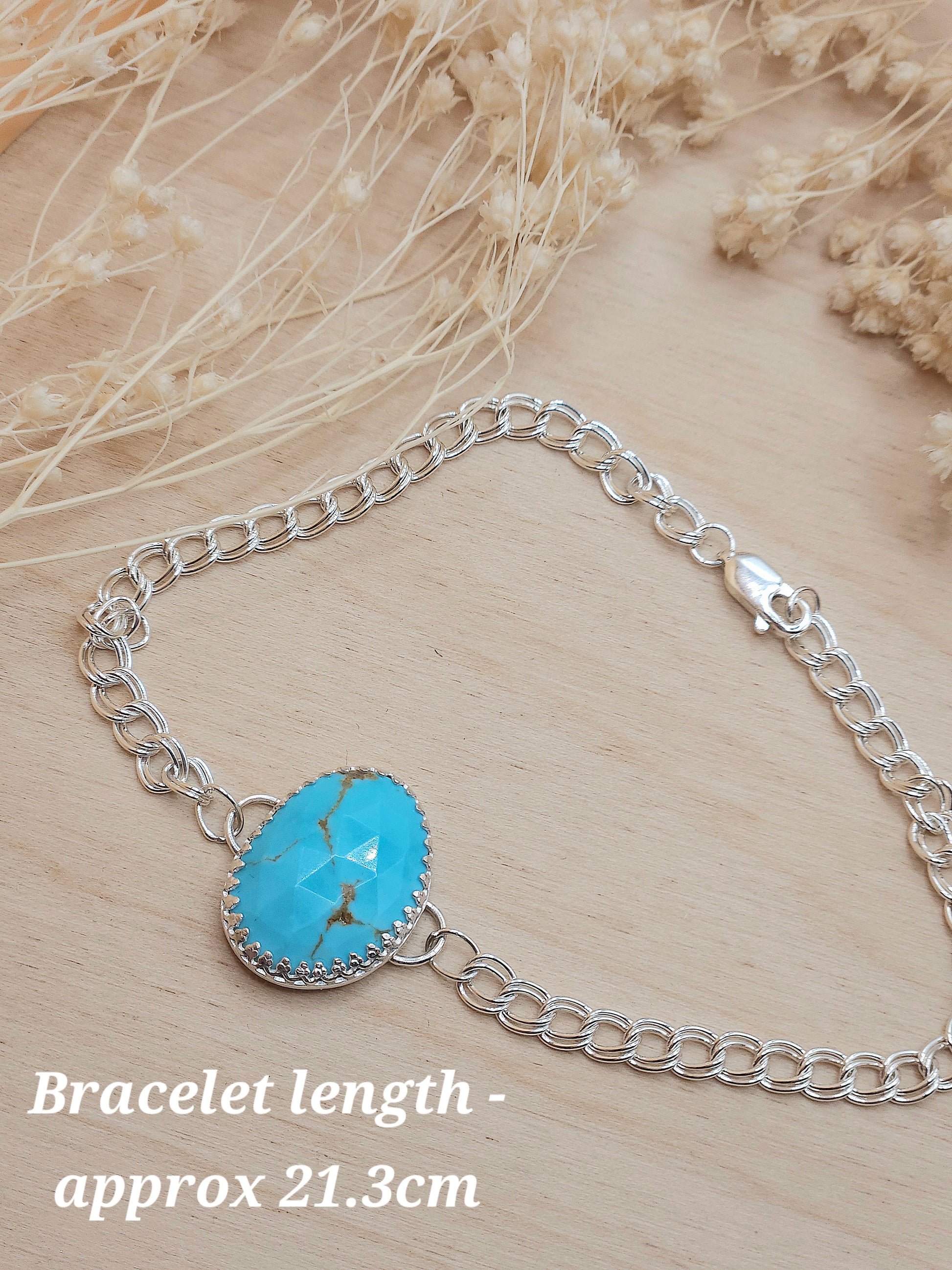 A silver chain bracelet with blue turquoise gemstone, displayed on a wooden background with dried flowers. A note in the corner of the photo reads 'Bracelet length - approx 21.3cm'.