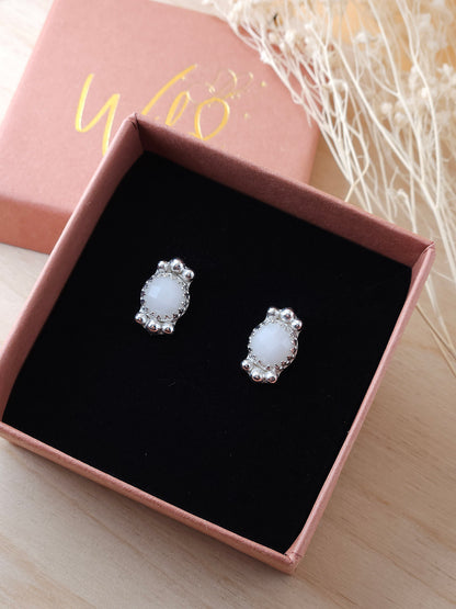 A pair of white agate silver stud earrings with silver embellishments displayed in an open gift box with dried flowers in the background.