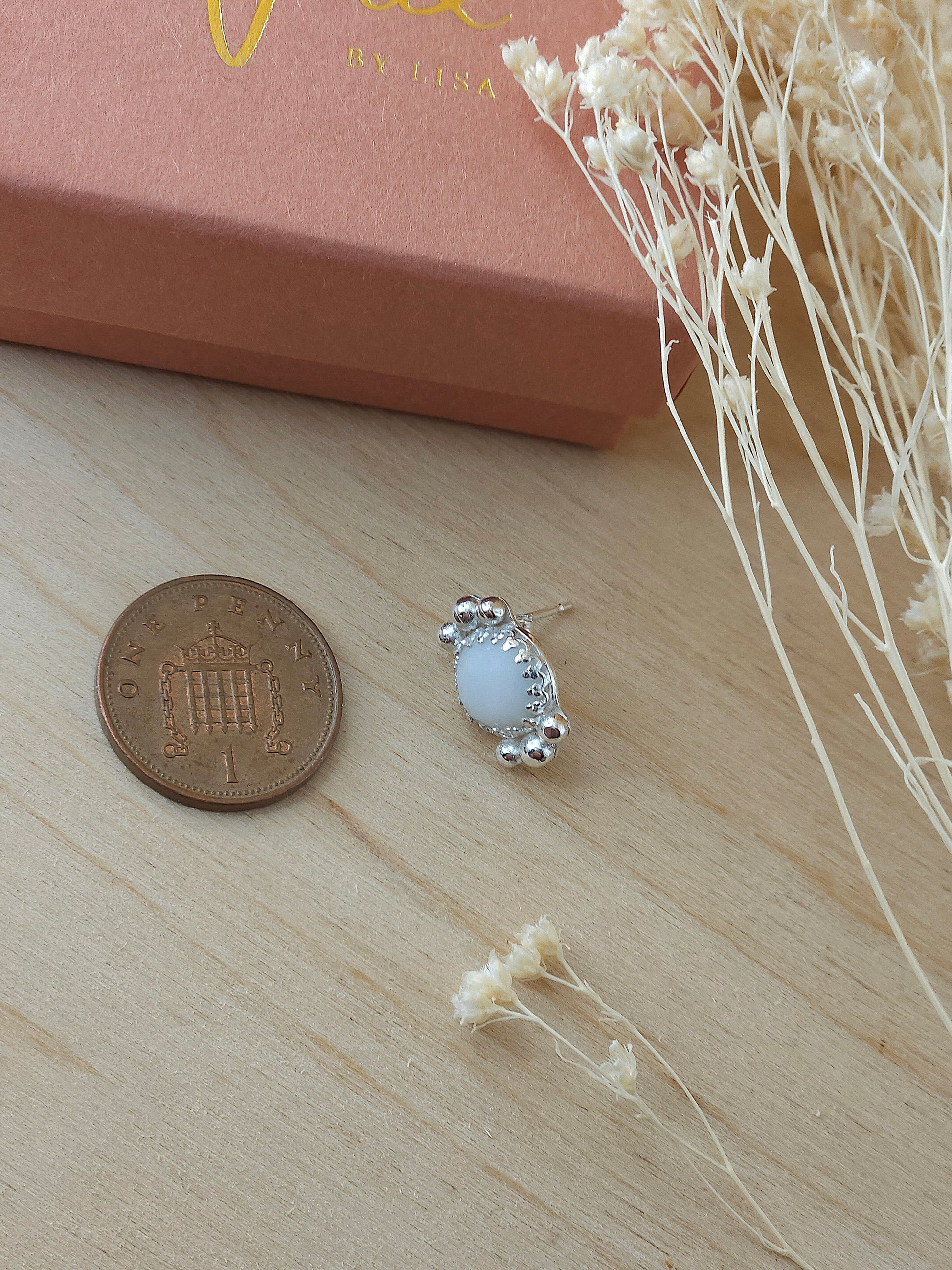 A single white agate silver stud earrings laying next to a one pence coin to show the size comparison. The earring is smaller than the coin.