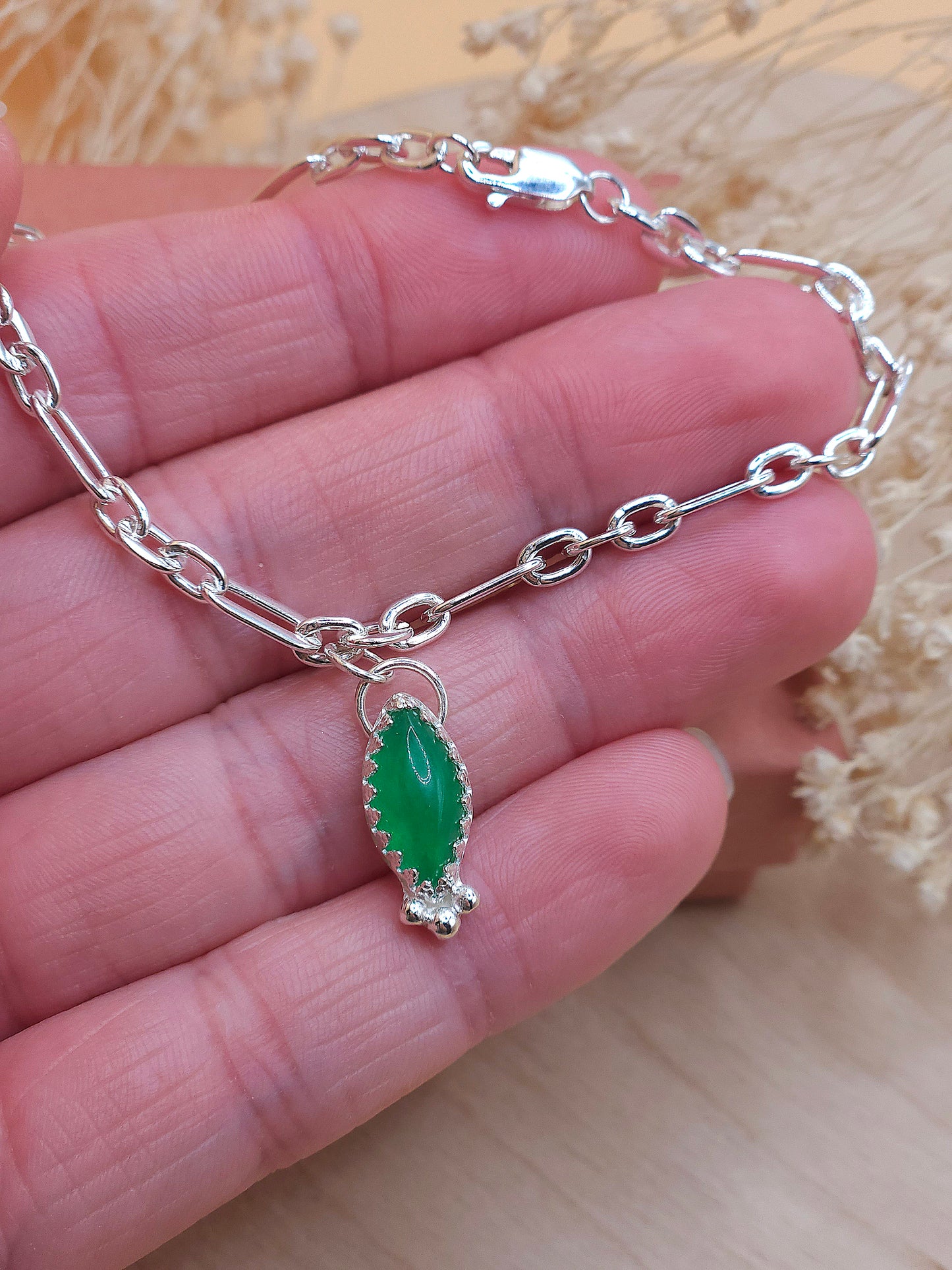 A hand holding a sterling silver link chain bracelet with a green aventurine gemstone charm. Dried flowers in the background.