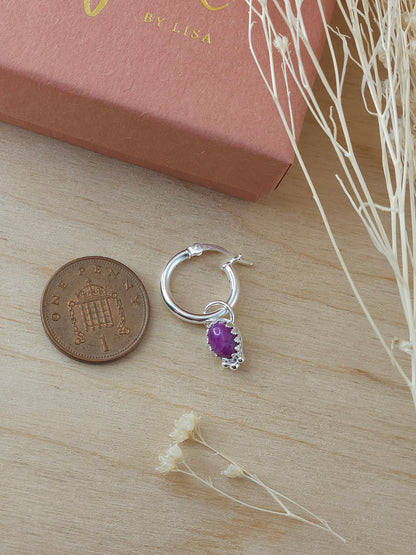A single silver mini hoop earring with ruby gemstone charm lay on a wooden surface next to a one pence coin.