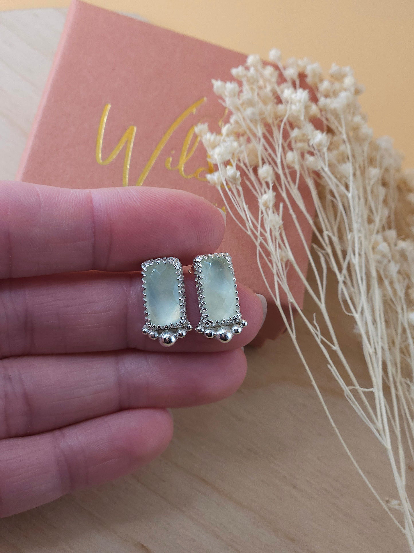 A hand holding a pair of silver stud earrings with green prehnite gemstones and silver embellishments.