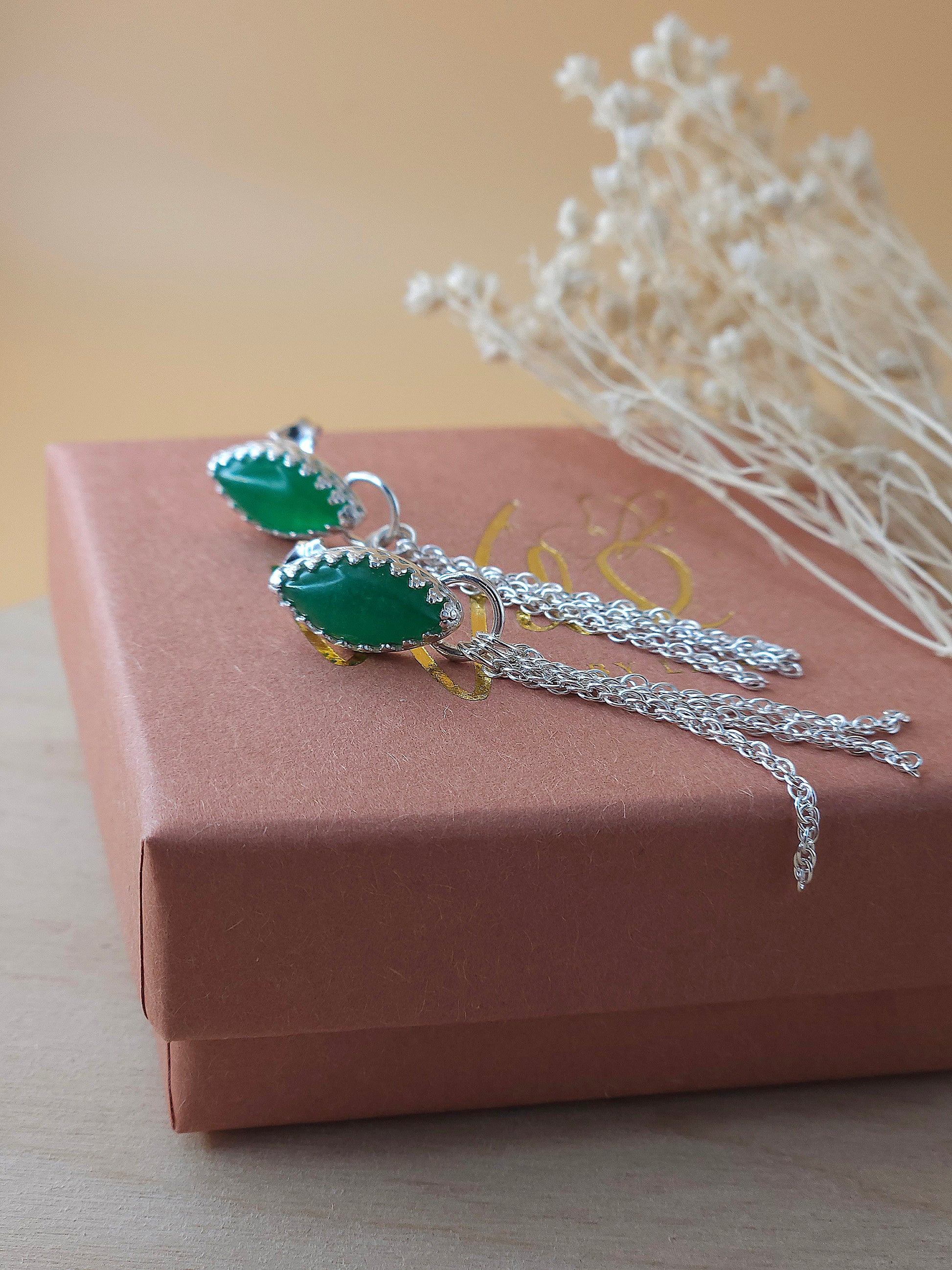 A pair of silver chain earrings with green aventurine gemstones laying on a gift box with dried flowers in the background.