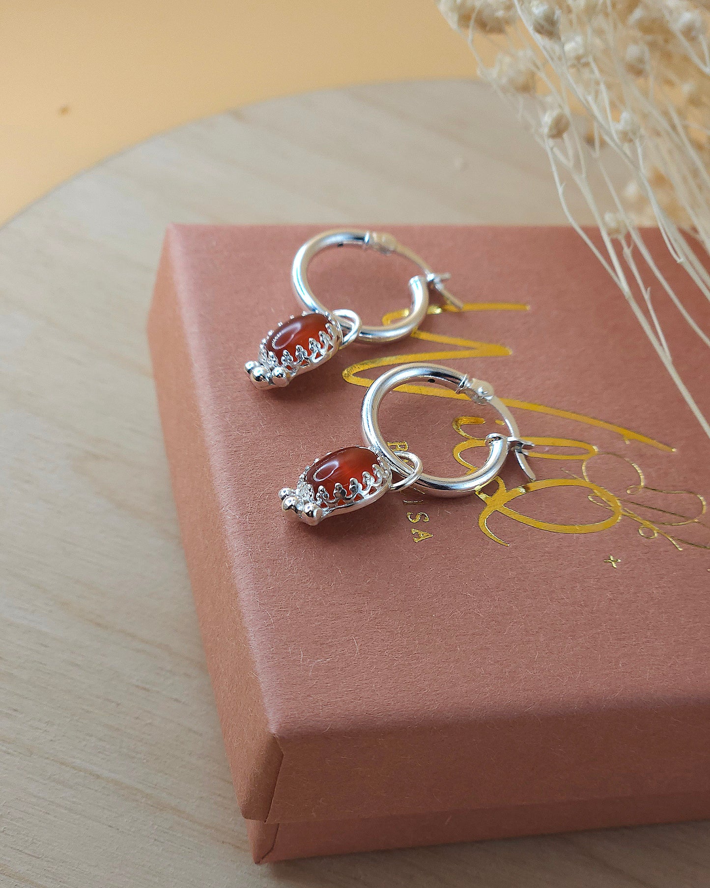 A pair of mini silver hoop earrings with red carnelian gemstone charms displayed on a terracotta and gold coloured gift box with dried flowers. Showing the side view of the earrings.