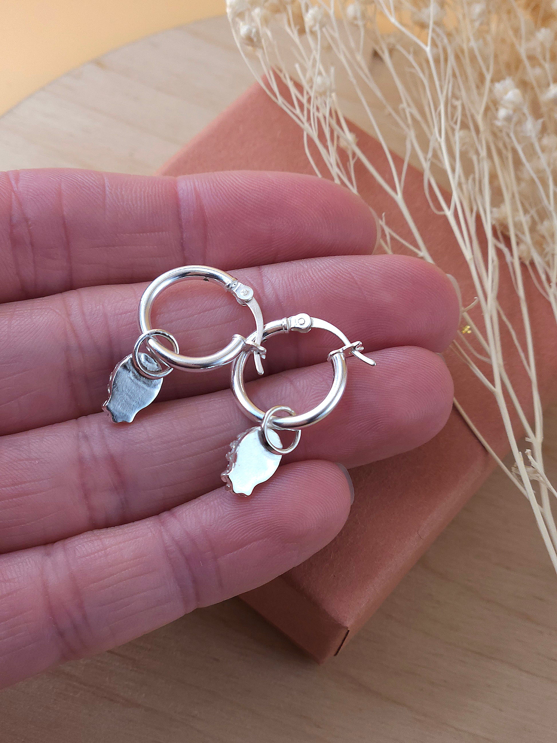 A hand holding a pair of mini silver hoop earrings showing the back of the gemstone setting.