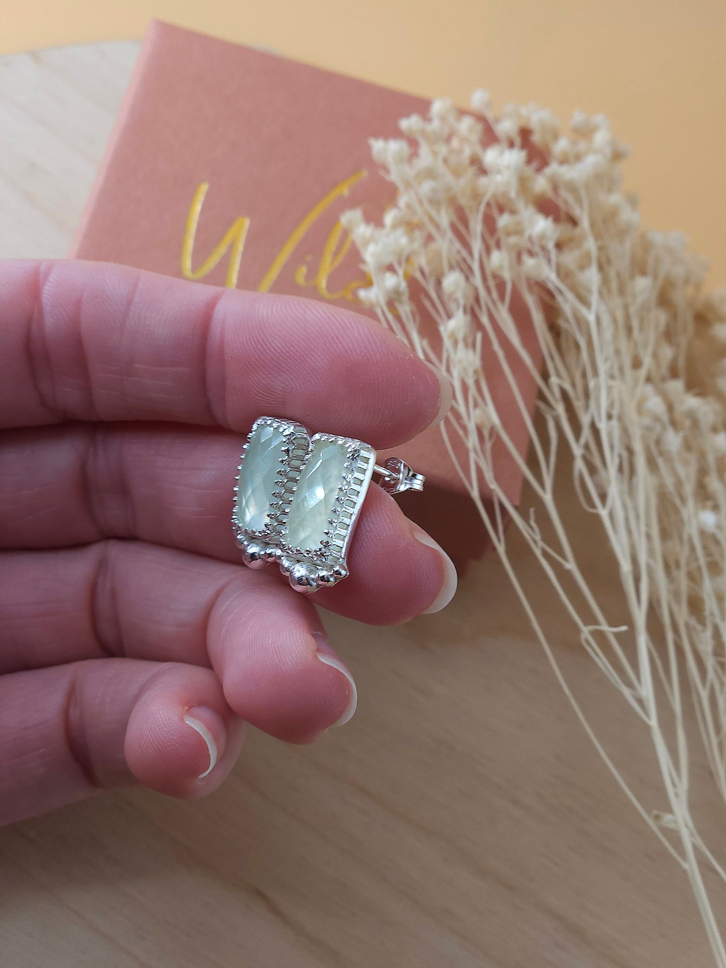 A hand holding a pair of sterling silver stud earrings with green prehnite gemstones and silver embellishments. Showing the side view of the earrings.