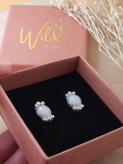 A hand holding an open gift box presenting a pair of sterling silver stud earrings with white agate gemstones and silver embellishments.